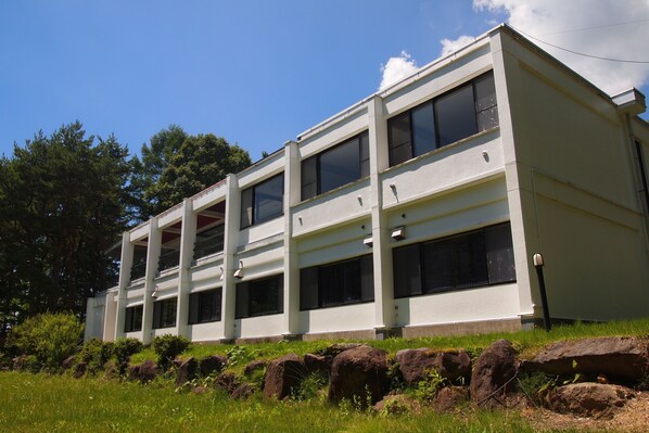 A modern building with multiple windows and a flat roof, surrounded by greenery and rocks.