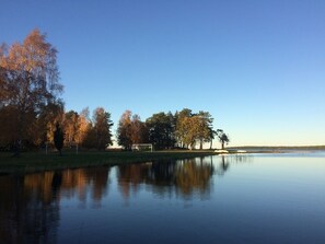 A calm lake with trees reflecting on the water, a grassy area, and a clear sky. - Pensionat Grankullavik (Byxelkrok)