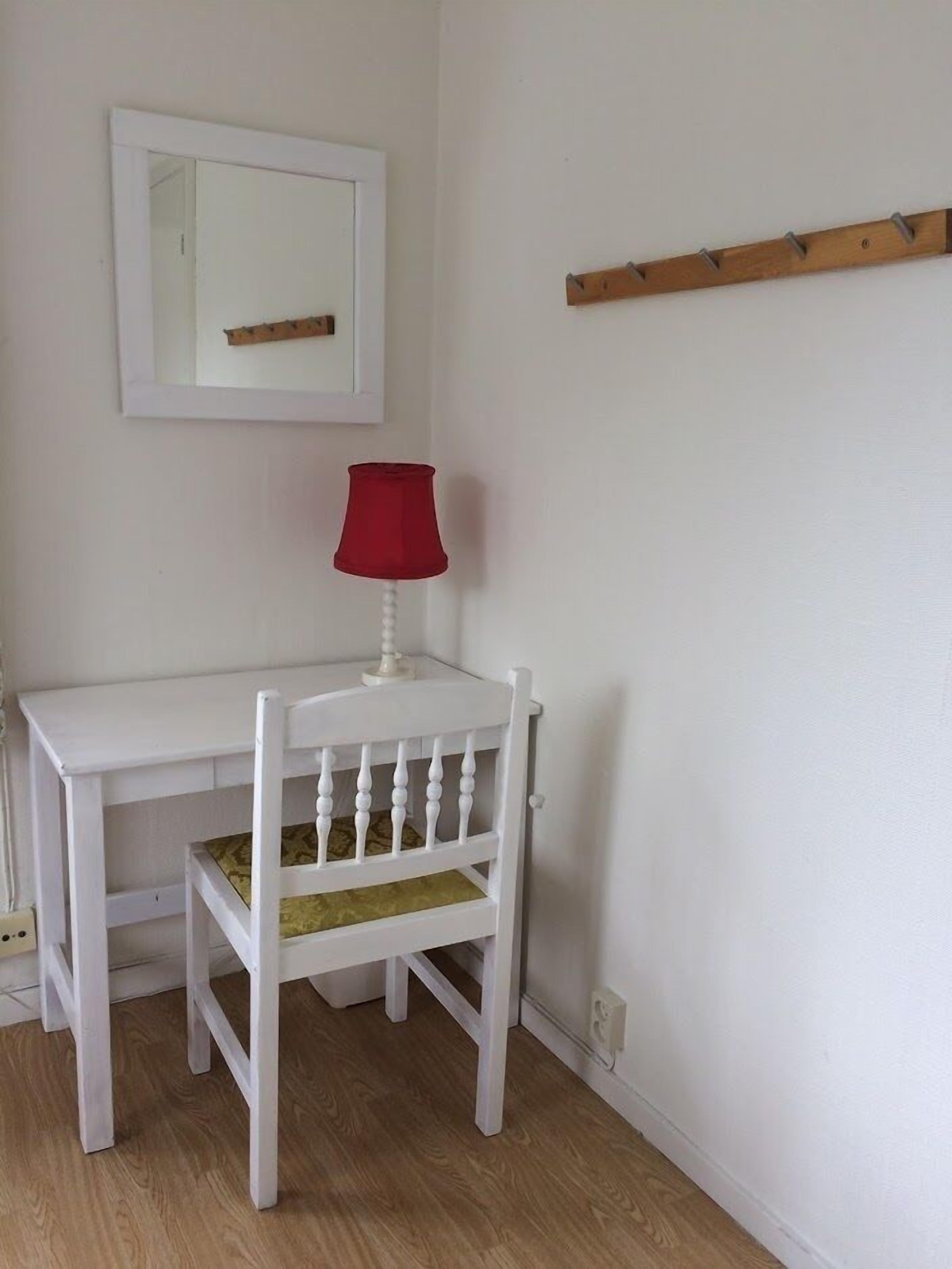 Ein weißer Schreibtisch mit einer roten Lampe und ein Stuhl mit einem grünen Kissen in einem Zimmer mit weißen Wänden und Holzboden.