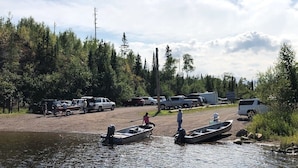 A campsite with vehicles and boats by a river, surrounded by trees.