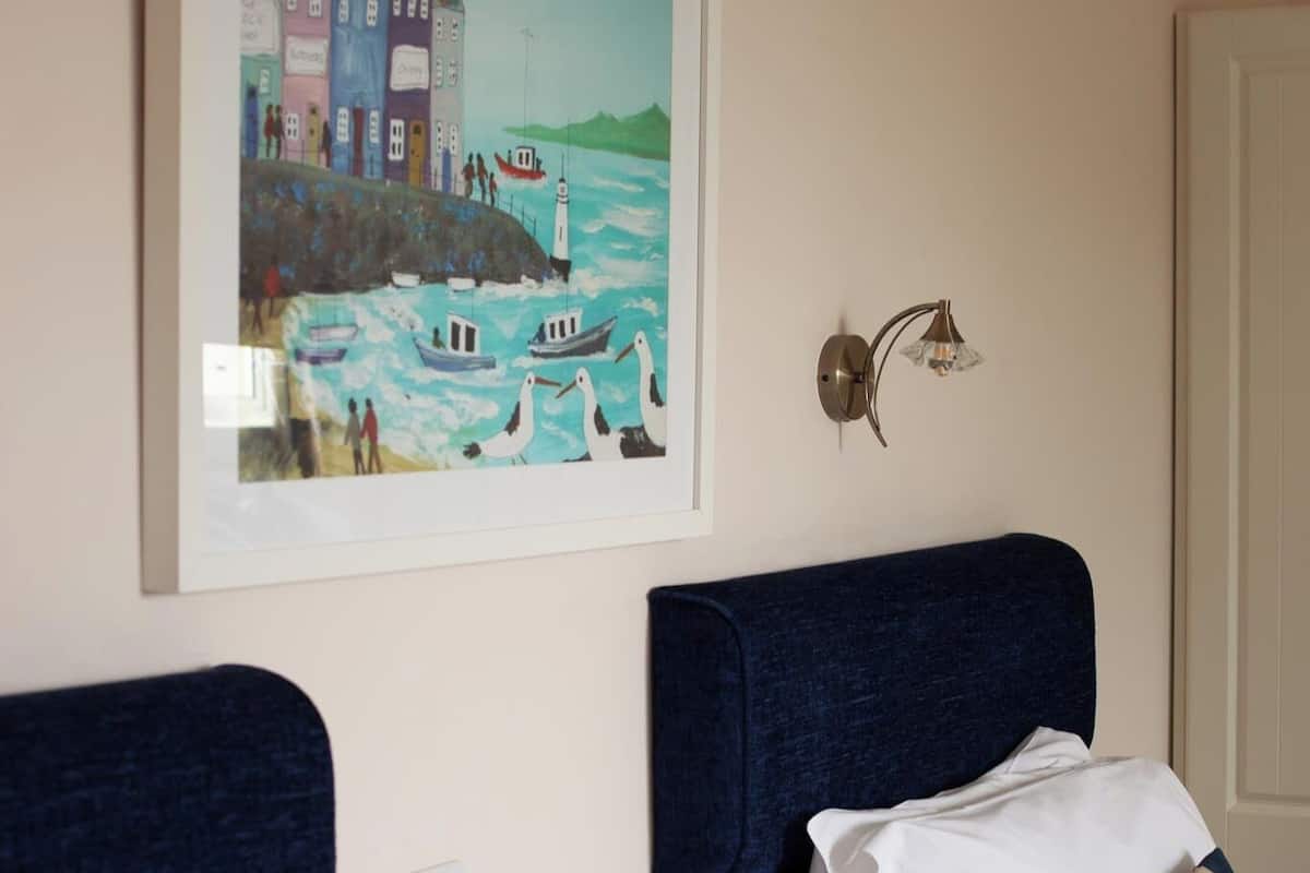 A hotel room with two beds, a framed painting of a coastal scene, a wall-mounted light fixture, and a door.