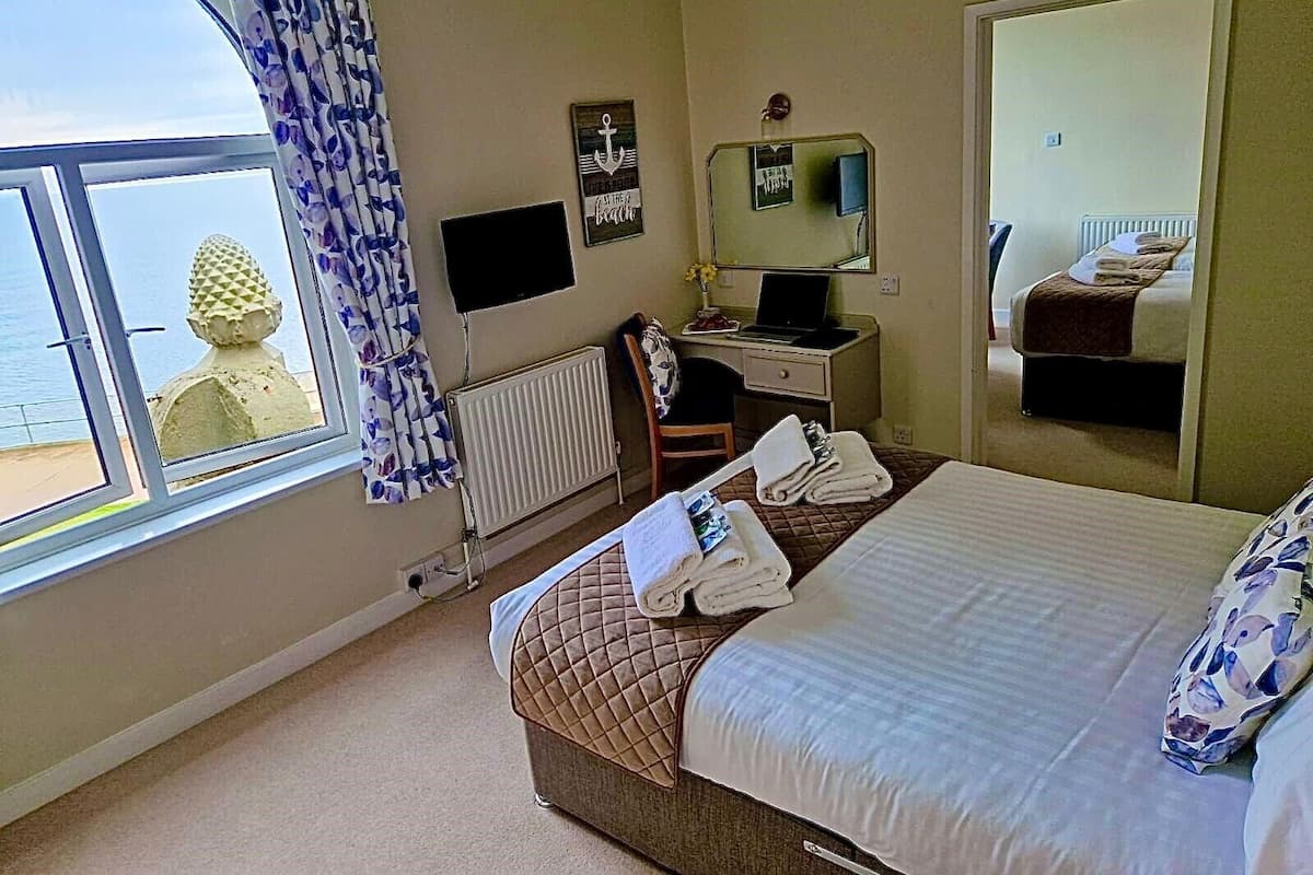 A hotel room with a bed, a desk with a laptop, a chair, a radiator, a window with curtains, and a view of the sea.