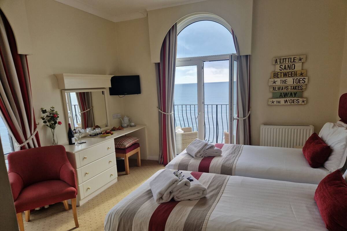 A bedroom with two beds, a desk, a chair, a TV, and a window with a view of the sea.