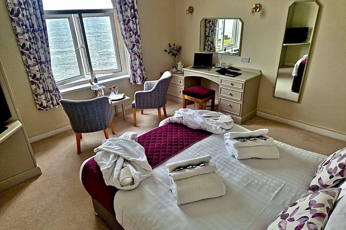 A hotel room with a bed, a desk with a laptop, two chairs, a small table, and a window with curtains.