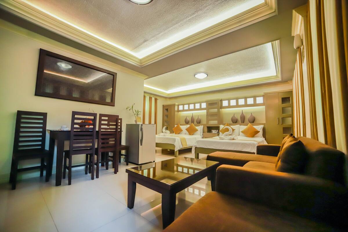 A hotel room with a dining area, a sofa, a bed with multiple pillows, and a mirror.