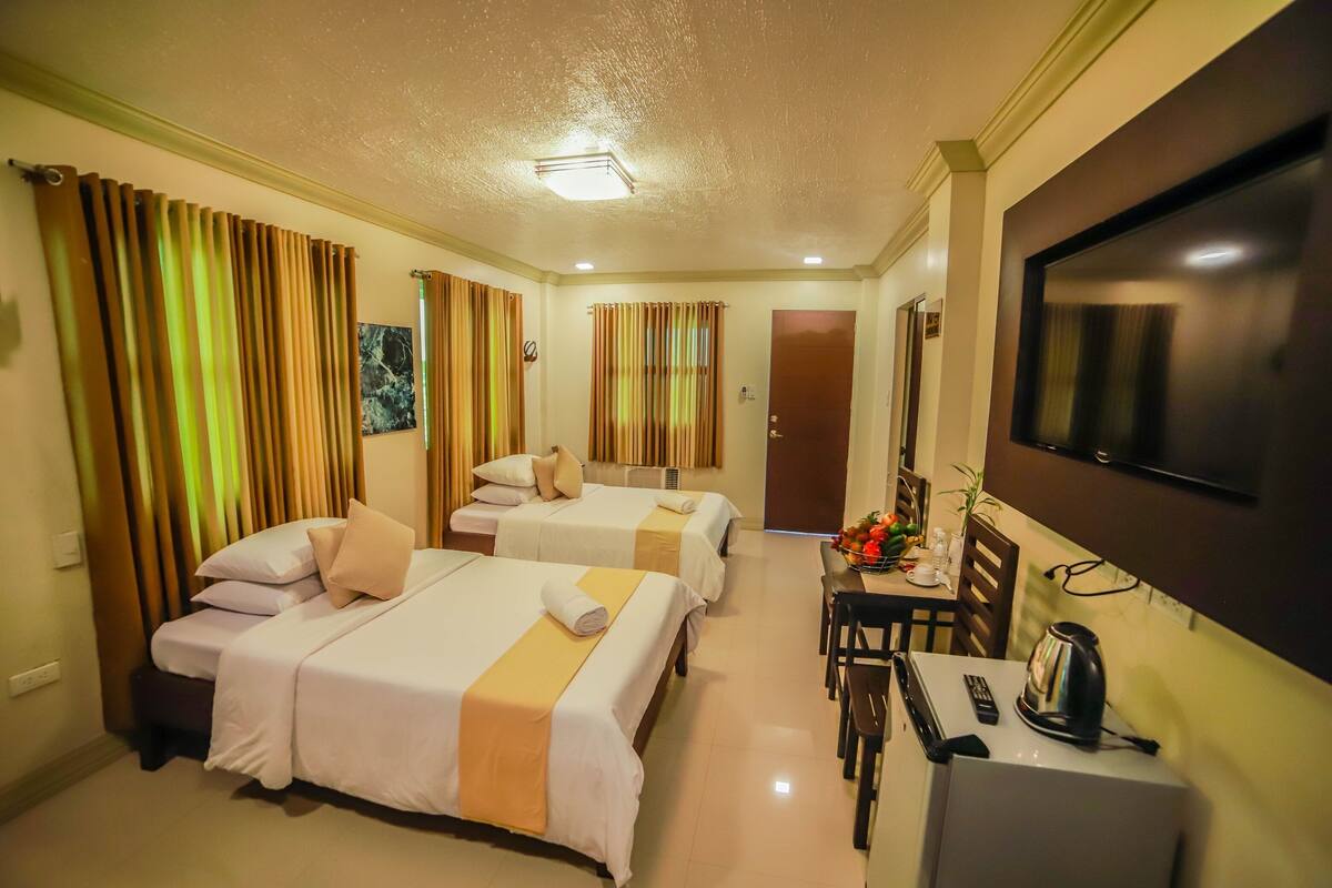 A hotel room with two beds, a desk with a kettle, a TV, and curtains.