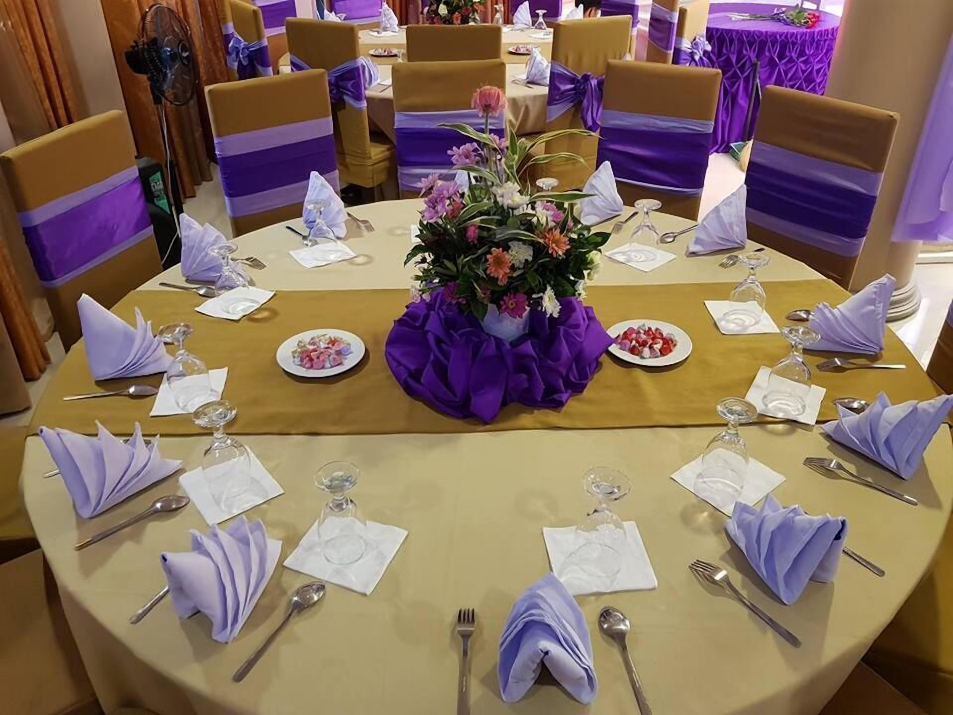 A round table set for a formal event with a central floral arrangement, surrounded by chairs with purple sashes, and a tablecloth with a gold border.