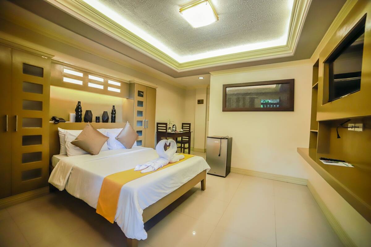 A hotel room with a bed, a desk, a refrigerator, and a TV mounted on the wall.