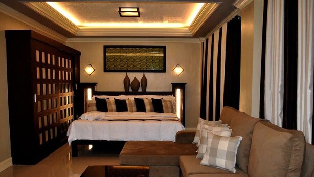 A bedroom with a large bed, a sofa, a wooden cabinet, and a decorative wall piece. - Villa Esmeralda Bryan's Resort Hotel and Restaurant (Palayan City)