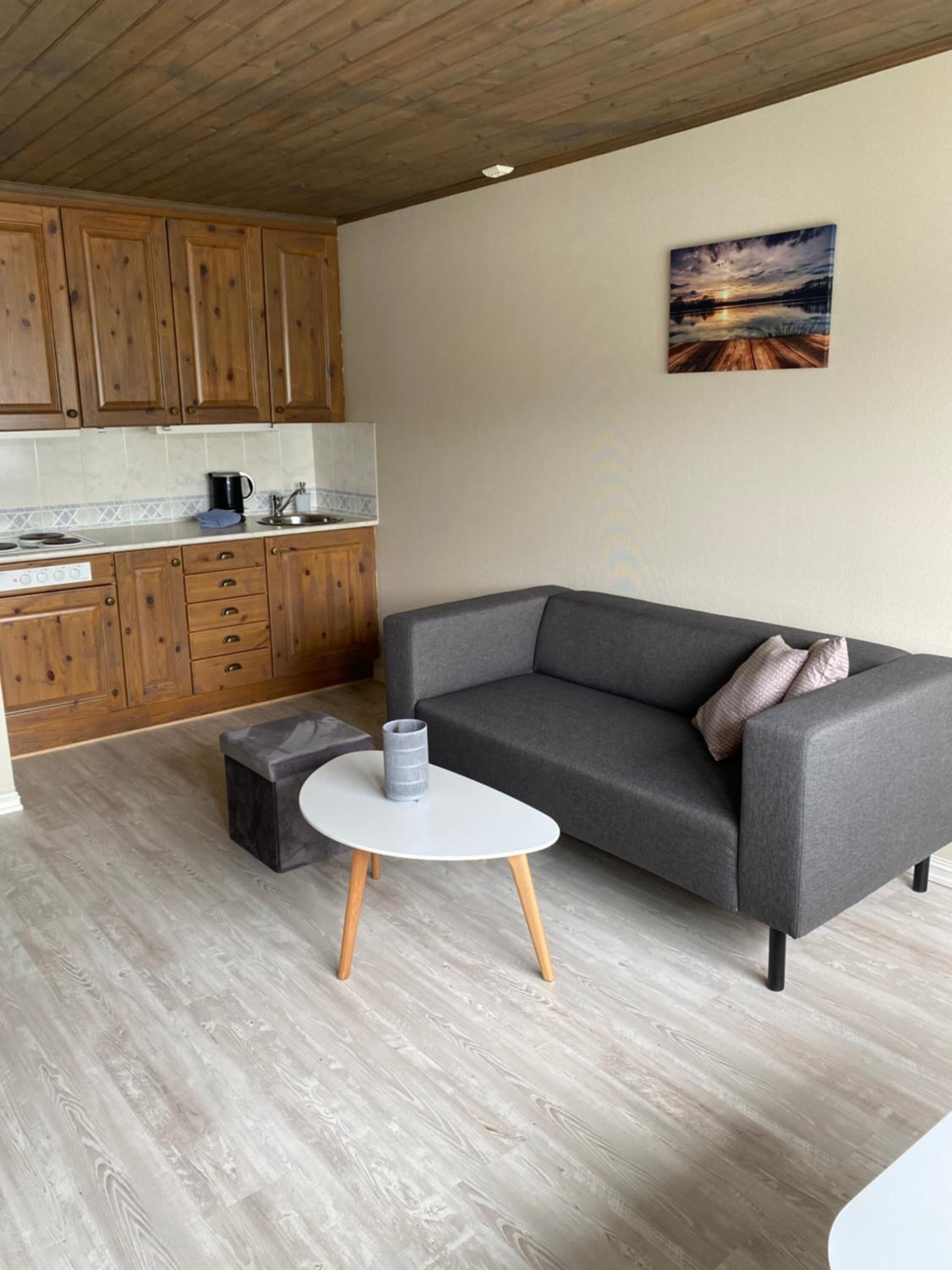 A compact living space with a grey sofa, a small round table, wooden cabinets, and a kitchen area.