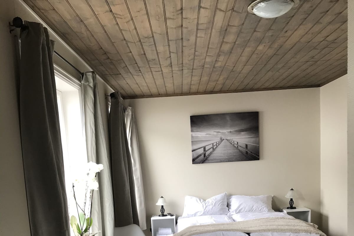A bedroom with a wooden ceiling, a bed with a gray bedspread, a desk with a chair, a nightstand with a lamp, and a window with curtains.
