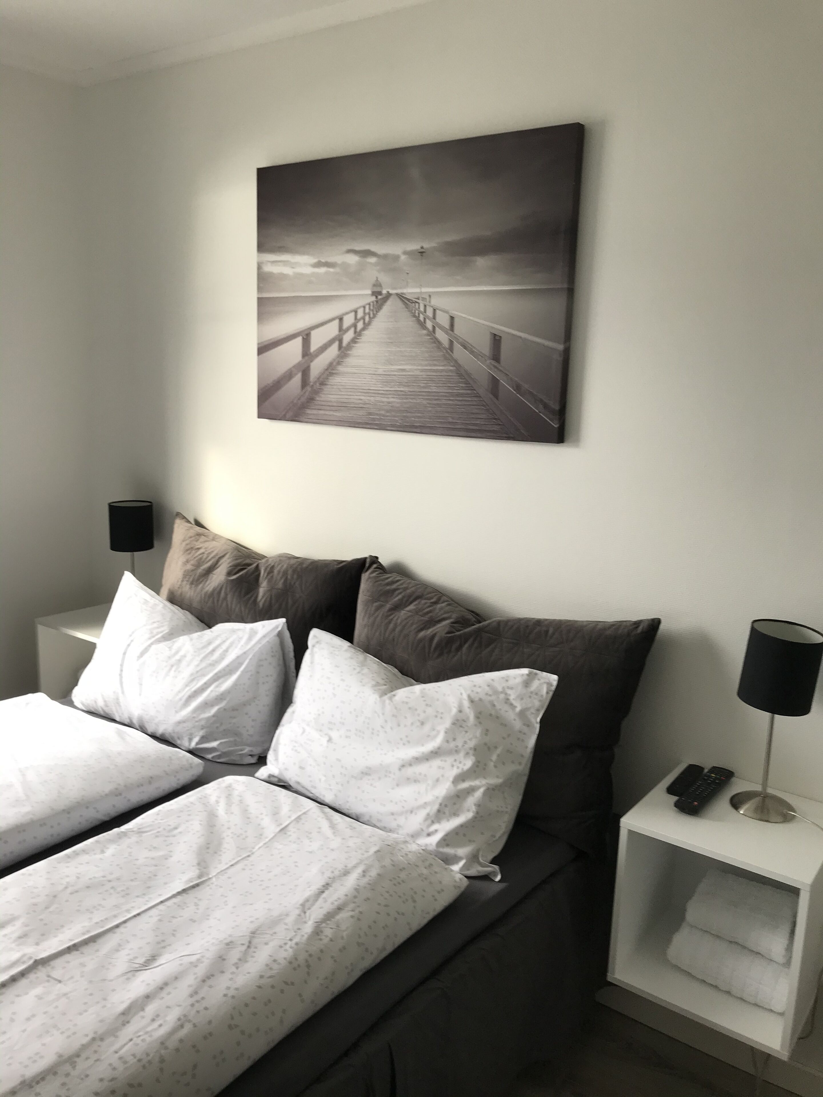 A bedroom with a bed, pillows, a nightstand, a lamp, and a wall-mounted picture of a pier.