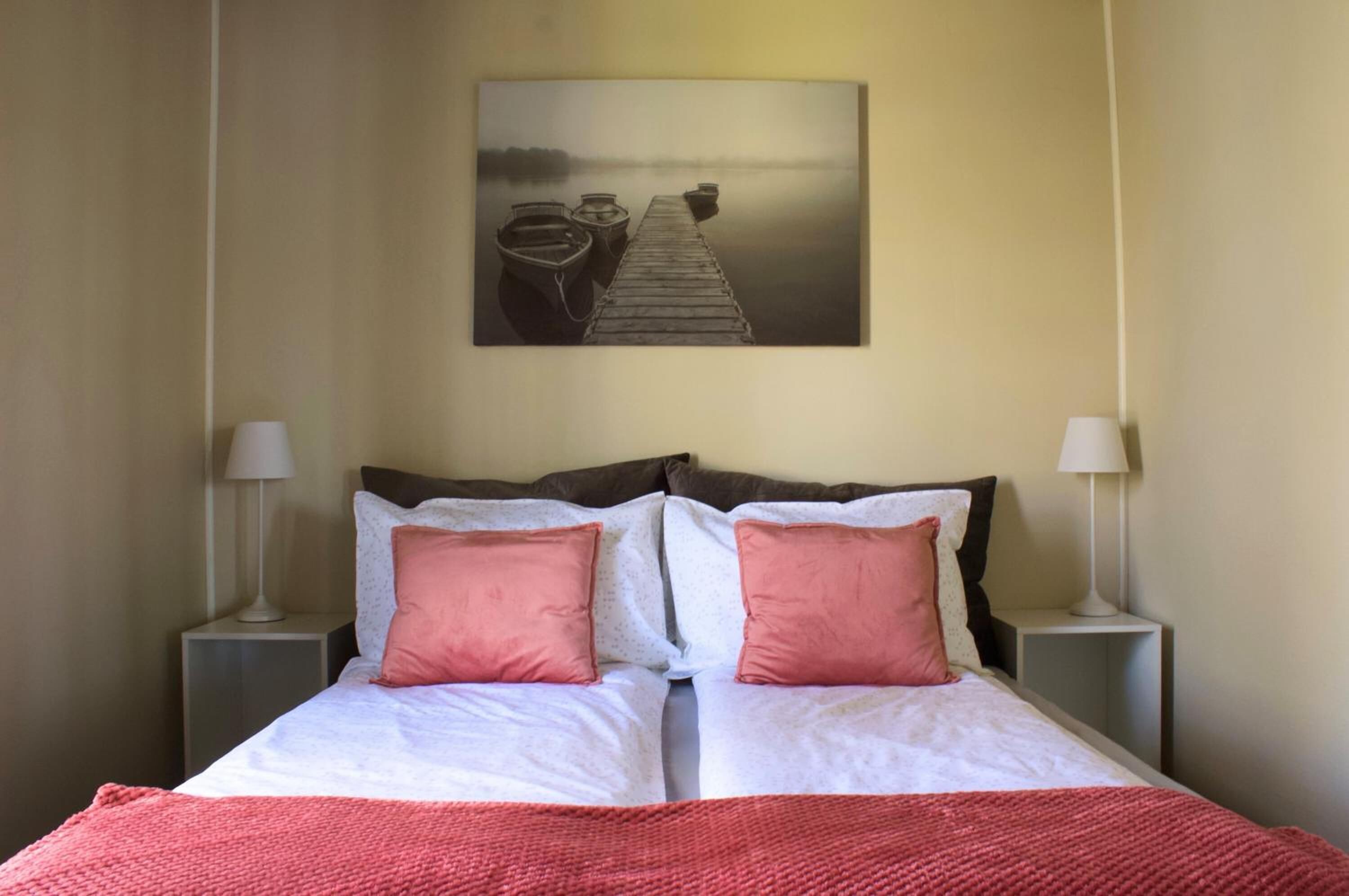Ein Doppelbett mit zwei Kissen, einer roten Decke und einem Gemälde von einem Steg mit Booten an der Wand darüber.