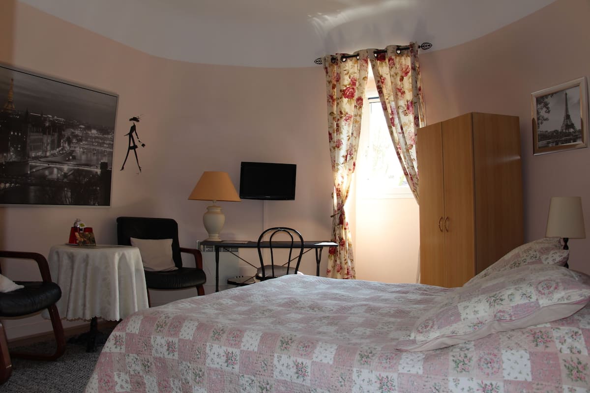 A bedroom with a bed, a desk with a chair, a television, a wardrobe, and a window with curtains.