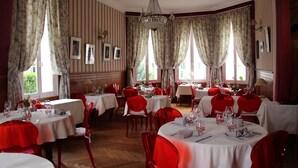 Un comedor con varias mesas y sillas, ventanas amplias con cortinas y una araña.