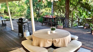 A round table with cushioned chairs, a grill, and potted plants on a wooden deck.