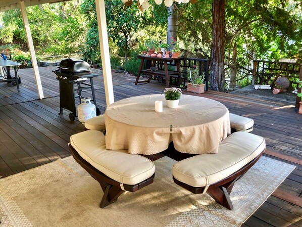 A round table with cushioned chairs, a grill, and potted plants on a wooden deck.