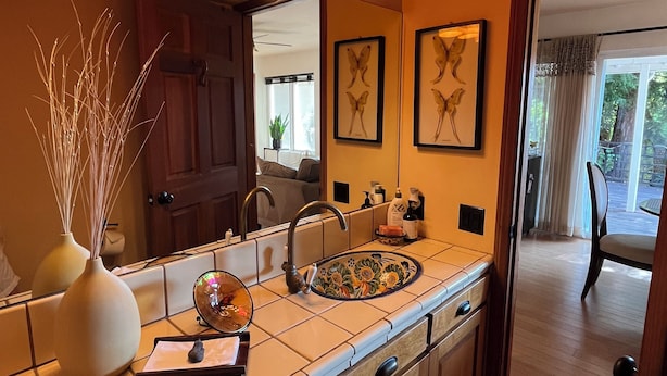 A modern kitchen with a large island, a double sink, and pendant lighting. There are framed butterfly art pieces on the wall and a view of the outdoor area through the door.