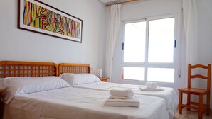 Une chambre d’hôtel avec deux lits, une tête de lit en bois, une grande fenêtre avec des rideaux, une chaise en bois et un tableau abstrait aux couleurs vives accroché au mur.