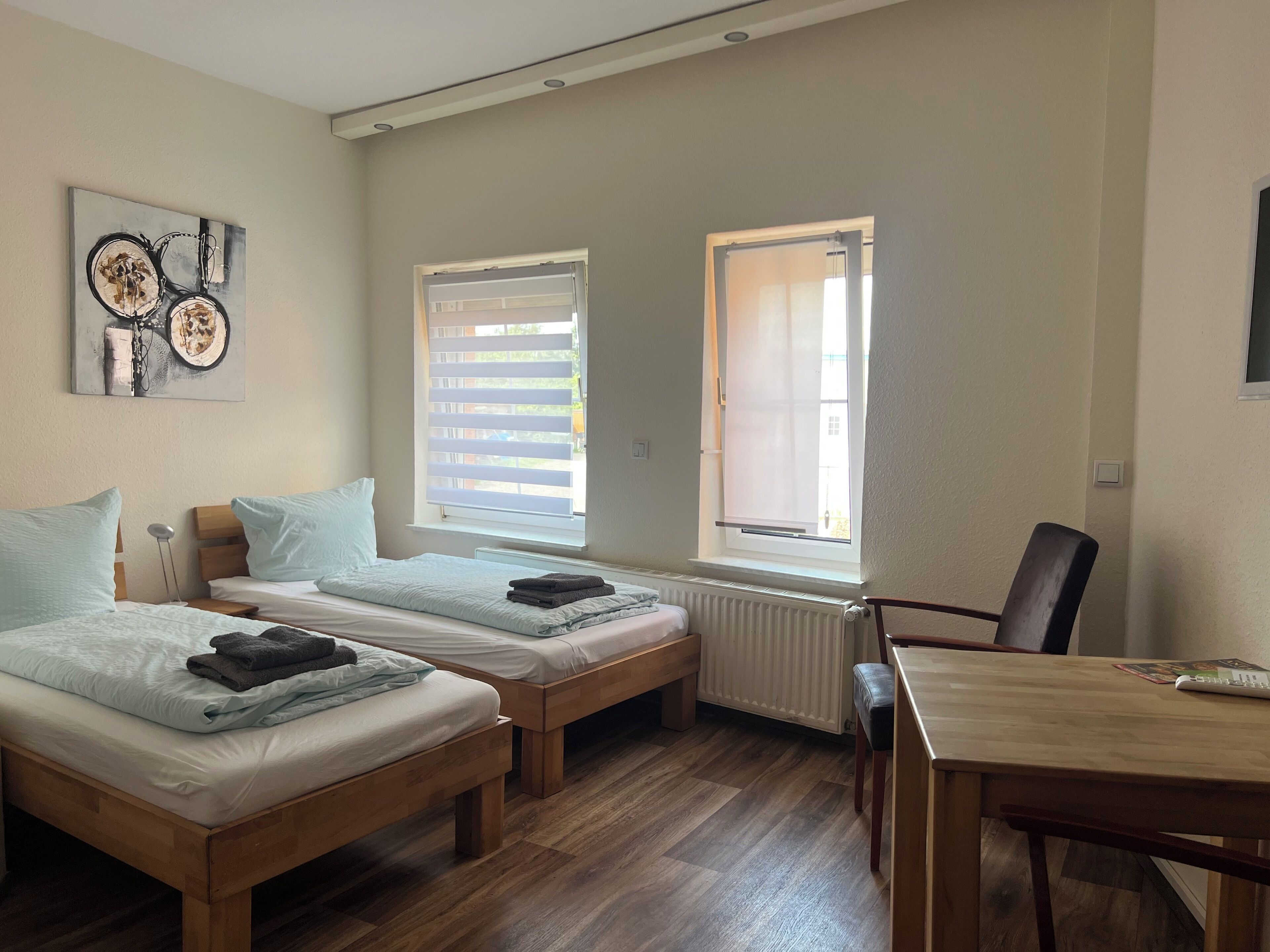 Una habitación con dos camas de madera, una mesa de madera y una silla. Hay dos ventanas con persianas, una de ellas abierta.