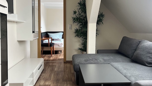 Ein modernes, kompaktes Wohnzimmer mit einer Couch, einem kleinen Tisch und einer integrierten Sitzbank. Eine große Pflanze sorgt für Grün, und der Raum verfügt über einen Holzboden.