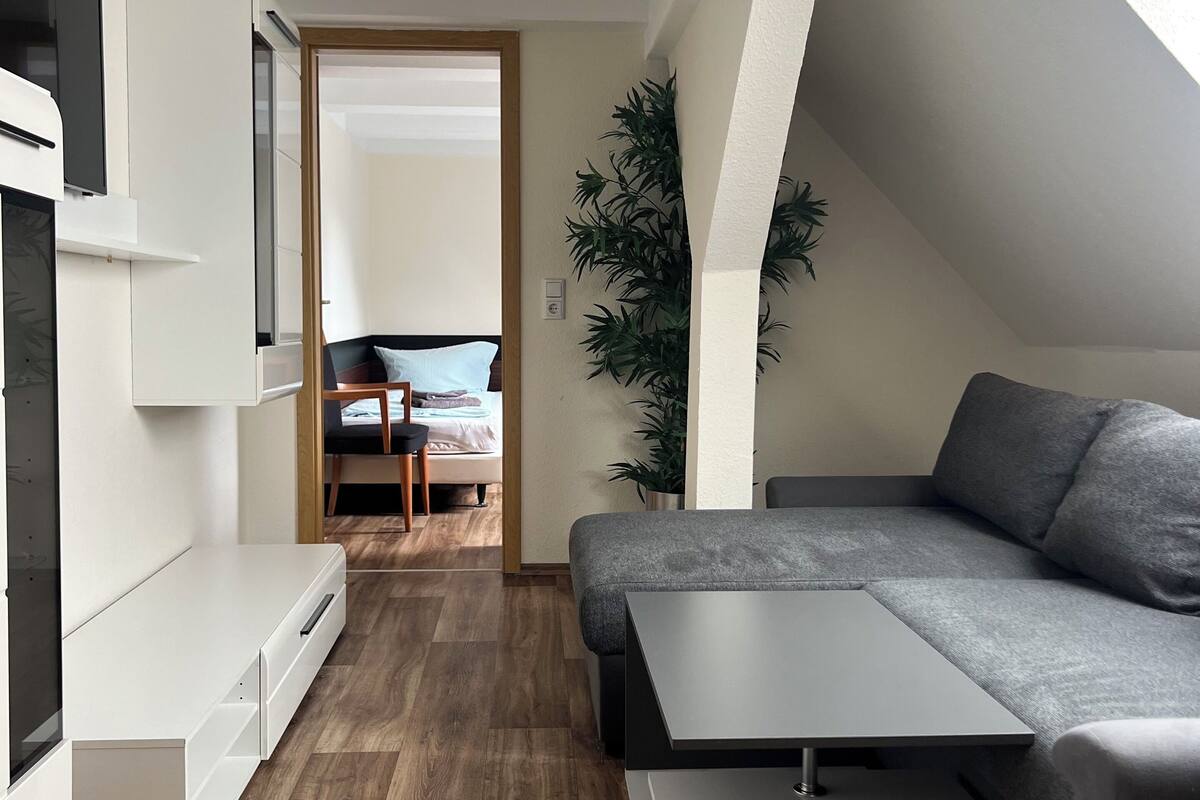 A modern, compact living space with a sofa, a small table, and a built-in bench. A large plant adds greenery, and the room has a wooden floor.