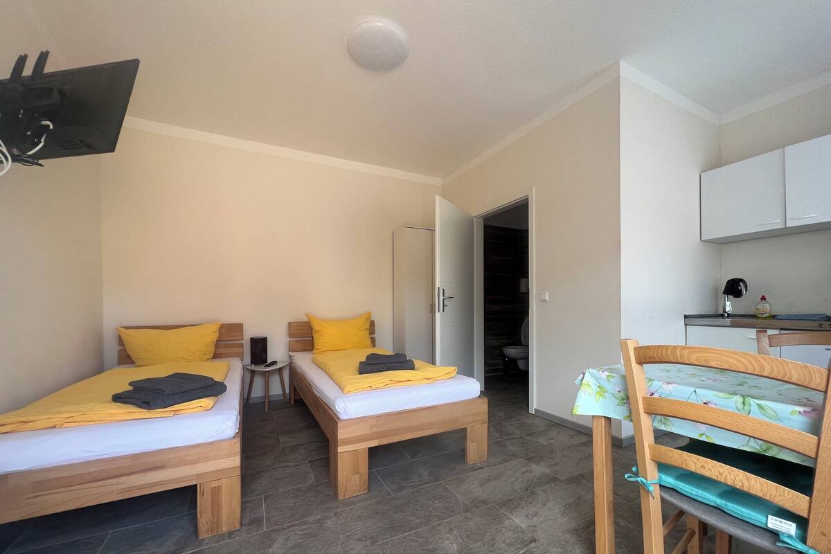 A room with two wooden beds, a small table, a chair, a TV mounted on the wall, and a kitchen area.