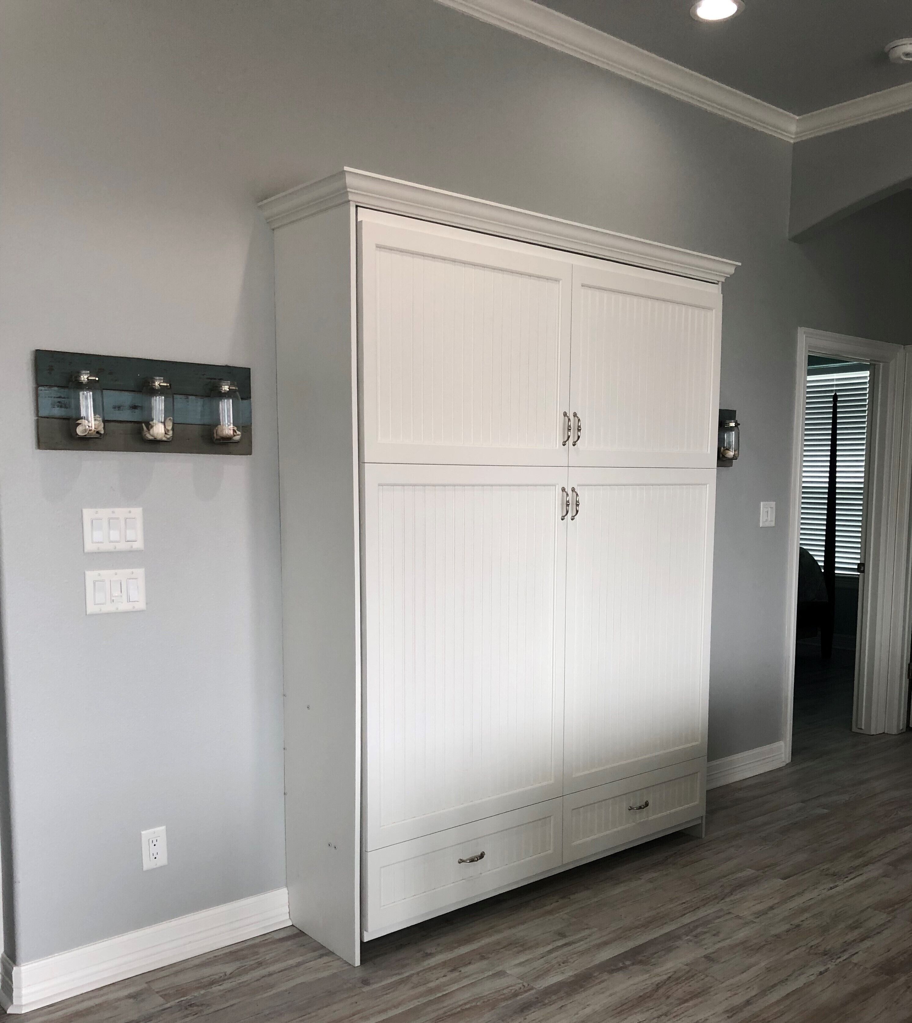 Une armoire blanche à deux portes et un tiroir, un interrupteur et une prise de courant sur le mur.
