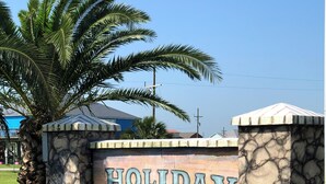 A stone wall with the words "HOLIDAY BEACH" painted on it, surrounded by grass and a palm tree. - Shark Shack ~ 3rd Row Home ~ Holiday Addition ~ Ocean View ~ Basketball Goal (Bolivar Peninsula)