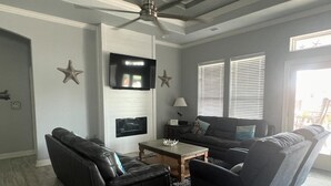 A living room with a ceiling fan, a flat-screen TV mounted on the wall, and a large window with blinds. - Shark Shack ~ 3rd Row Home ~ Holiday Addition ~ Ocean View ~ Basketball Goal (Bolivar Peninsula)