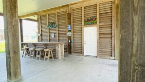 A covered outdoor area with wooden beams and a concrete floor, featuring a wooden table and stools, a door, and a sign with palm tree decorations. - Shark Shack ~ 3rd Row Home ~ Holiday Addition ~ Ocean View ~ Basketball Goal (Bolivar Peninsula)
