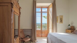 A bedroom with a bed, a chair, a nightstand, a mirror, and a view of the outdoors.