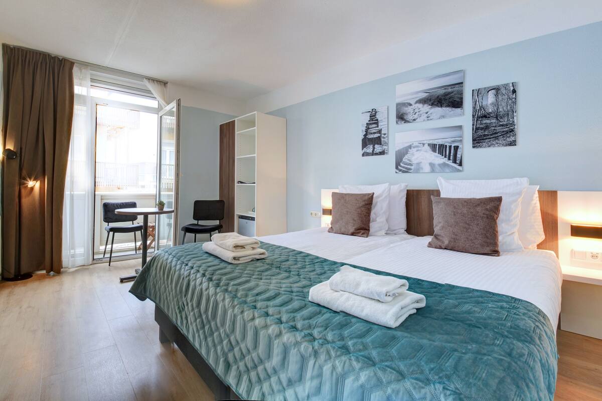 Ein Hotelzimmer mit einem großen Bett, einem Schreibtisch und einem Sessel. Drei Wandleuchten und ein Fenster mit Vorhängen sind vorhanden.