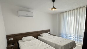 Habitación de hotel con dos camas, ventilador de techo, aire acondicionado, mesita de noche y ventana con cortinas translúcidas.