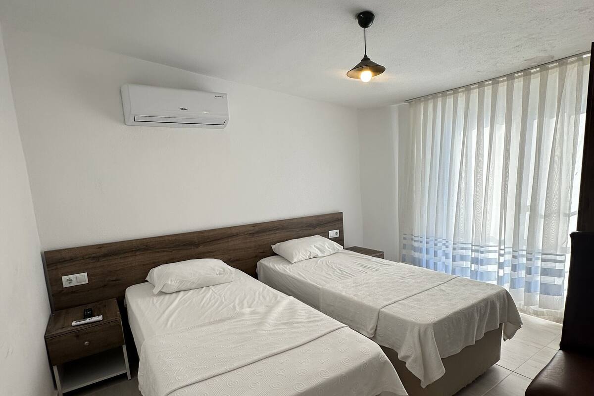 A hotel room with two beds, a ceiling fan, an air conditioning unit, a nightstand, and a window with sheer curtains.