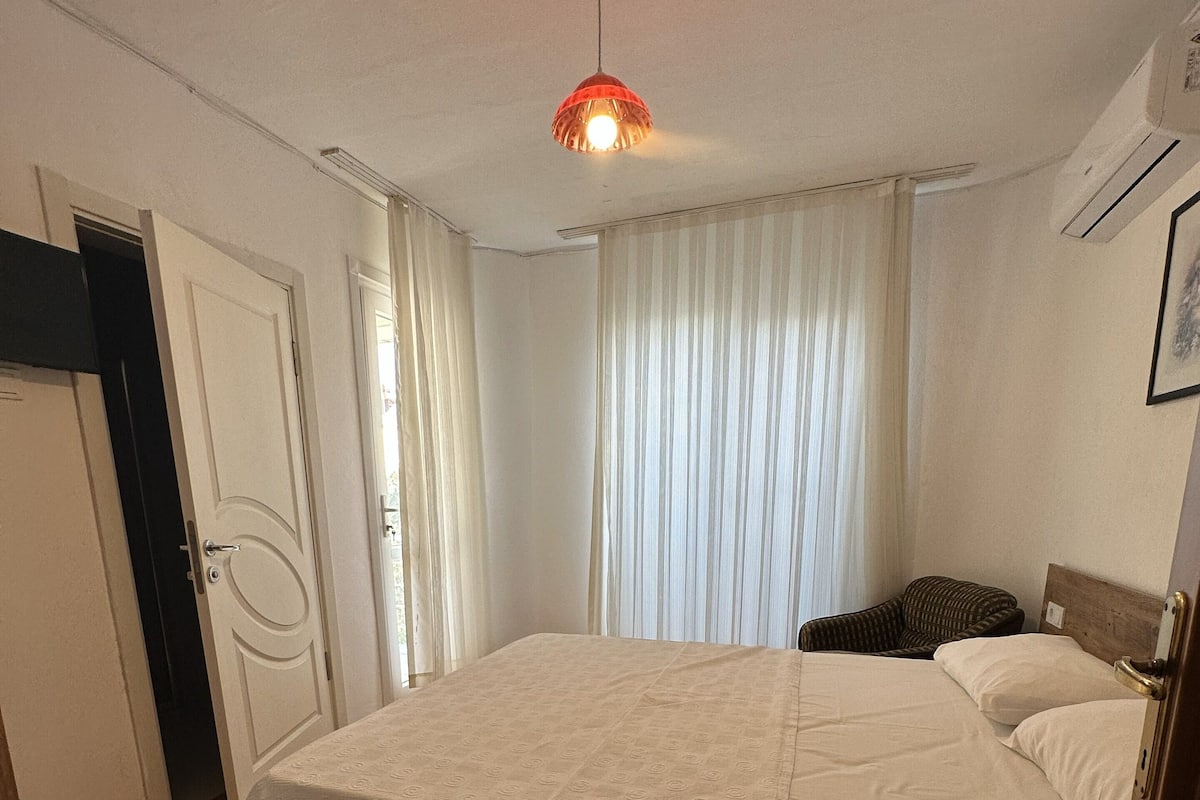 A bedroom with a large bed, a bedside table, a chair, a door, and a window with curtains.