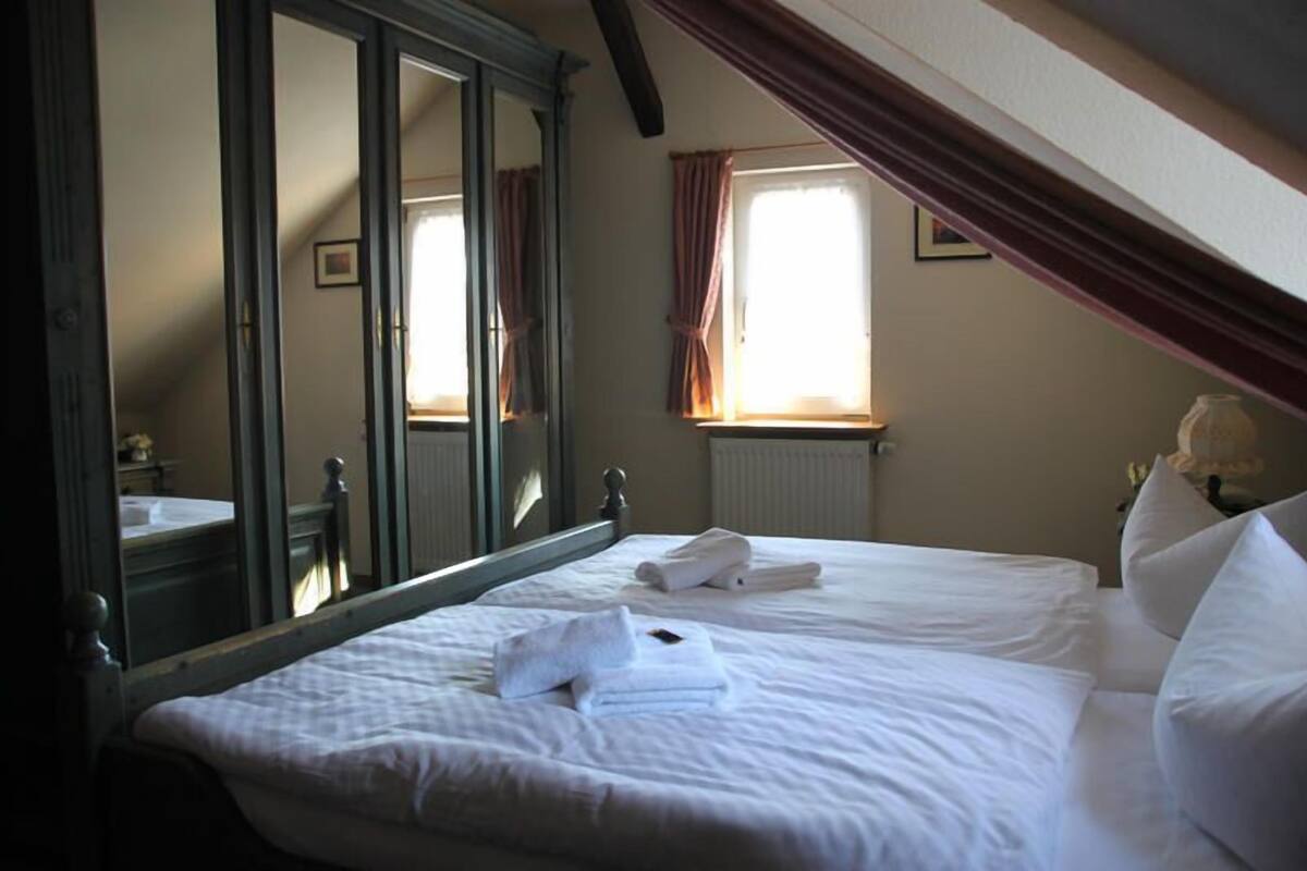 A bedroom with a large mirror, a bed with white linens, and a window with curtains.