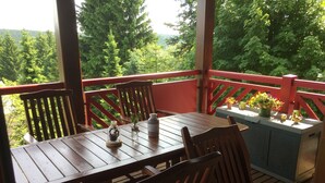 Eine Holzdiele mit Tisch und Stühlen, umgeben von einem roten Geländer, mit Blick auf einen bewaldeten Bereich.