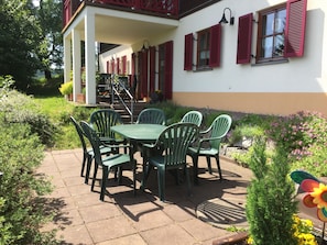 An outdoor patio with a table and chairs, surrounded by greenery and a building with red shutters.