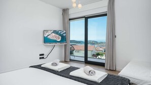 A modern hotel room with a bed, a TV mounted on the wall, and a large window offering a view of a coastal town.