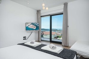 Une chambre d’hôtel moderne, équipée d’un lit, d’un téléviseur fixé au mur et d’une grande fenêtre donnant sur une ville côtière.