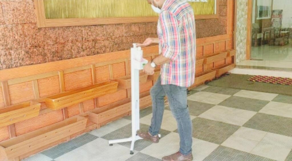 A man standing next to a hand sanitizer dispenser in a room with wooden benches and a checkered floor.