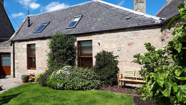 A stone cottage with a sloped roof, two windows, a wooden bench, and a garden with various plants. - North End Nairn The Cottage (Nairn)