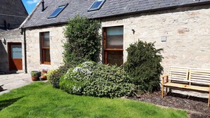 A stone building with a sloped roof, a wooden bench, and a small garden area with potted plants. - North End Nairn The Cottage (Nairn)