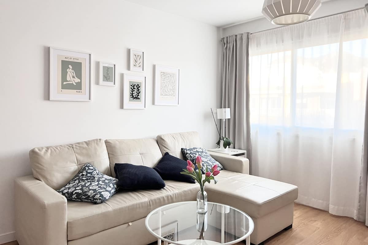 Una sala de estar moderna con un sofá color beige, una mesa de centro de vidrio y un florero con flores. Hay obras de arte enmarcadas en la pared y una lámpara sobre una mesita.
