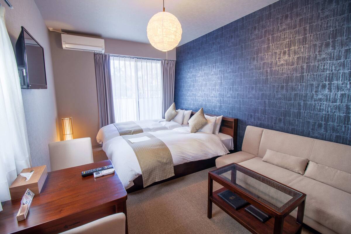 A hotel room with a large bed, a sofa, a wooden table, and a glass coffee table. There is a lamp, a TV mounted on the wall, and an air conditioning unit.