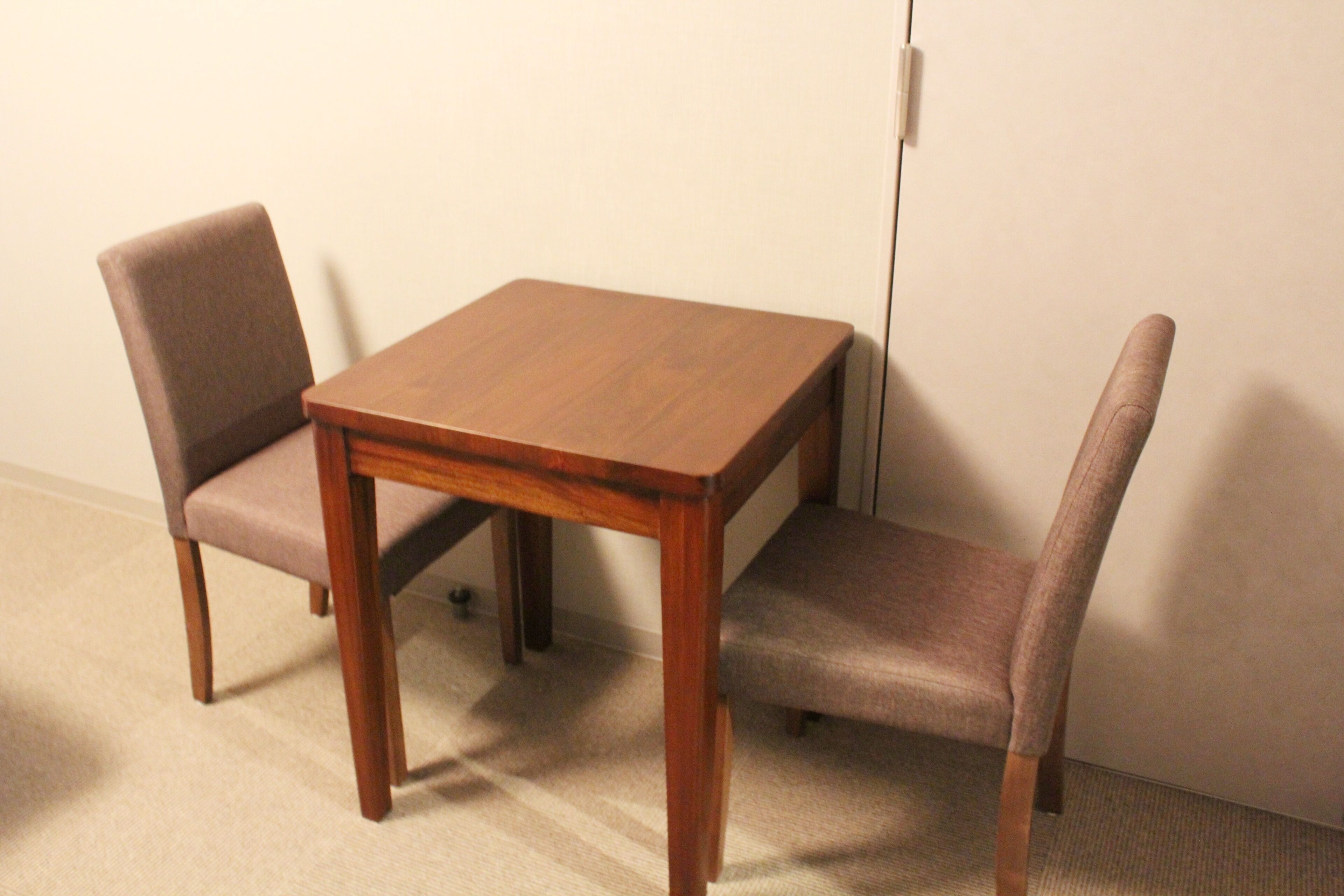 Une petite table en bois, deux chaises, un mur sobre et un sol recouvert d’une moquette.