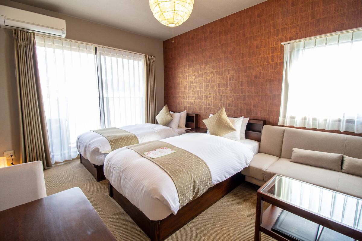 A hotel room with two beds, a sofa, a glass coffee table, and a brick wall.