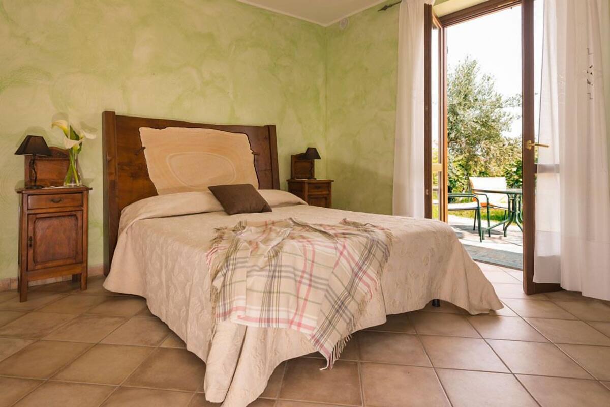 A bedroom with a large bed, two bedside tables, a nightstand, a window with curtains, and a view of the outdoors.