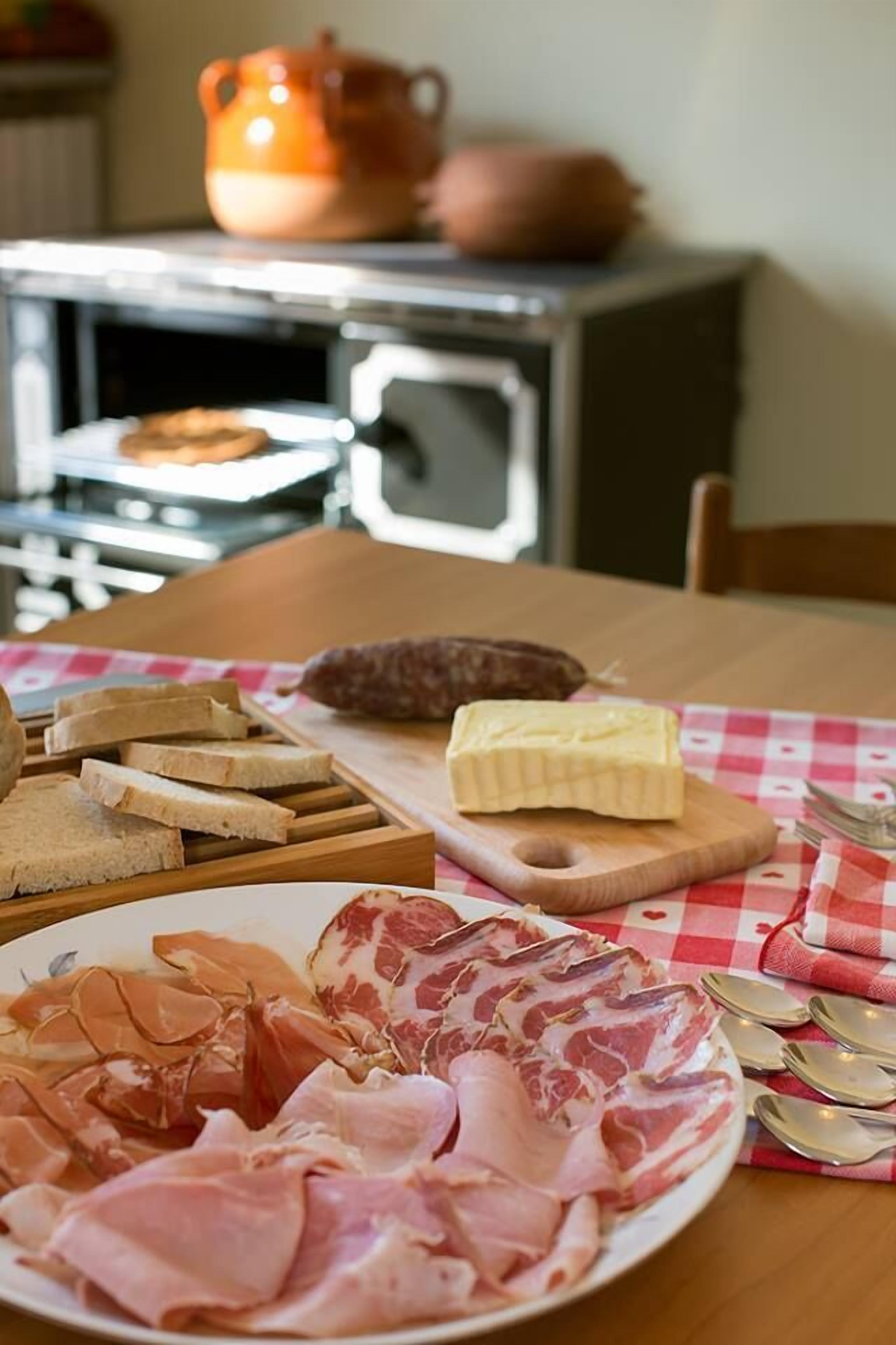 A table with various food items including bread, cheese, and meats, with a checkered tablecloth and a rustic kitchen in the background.